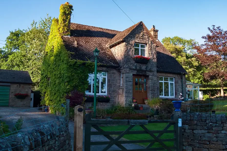 Bronte Cottage is illuminated by the evening sun. The cottage sits in a neatly kept garden behind a drystone wall. An old fashioned lamp-post stands in front of the cottage, inside the garden.