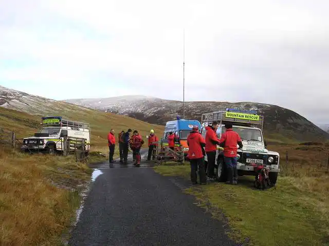 A mountain rescue team in operation. Two Land Rover Defender vehicles and a Transit van with a large radio mast park beside a lane in the mountain